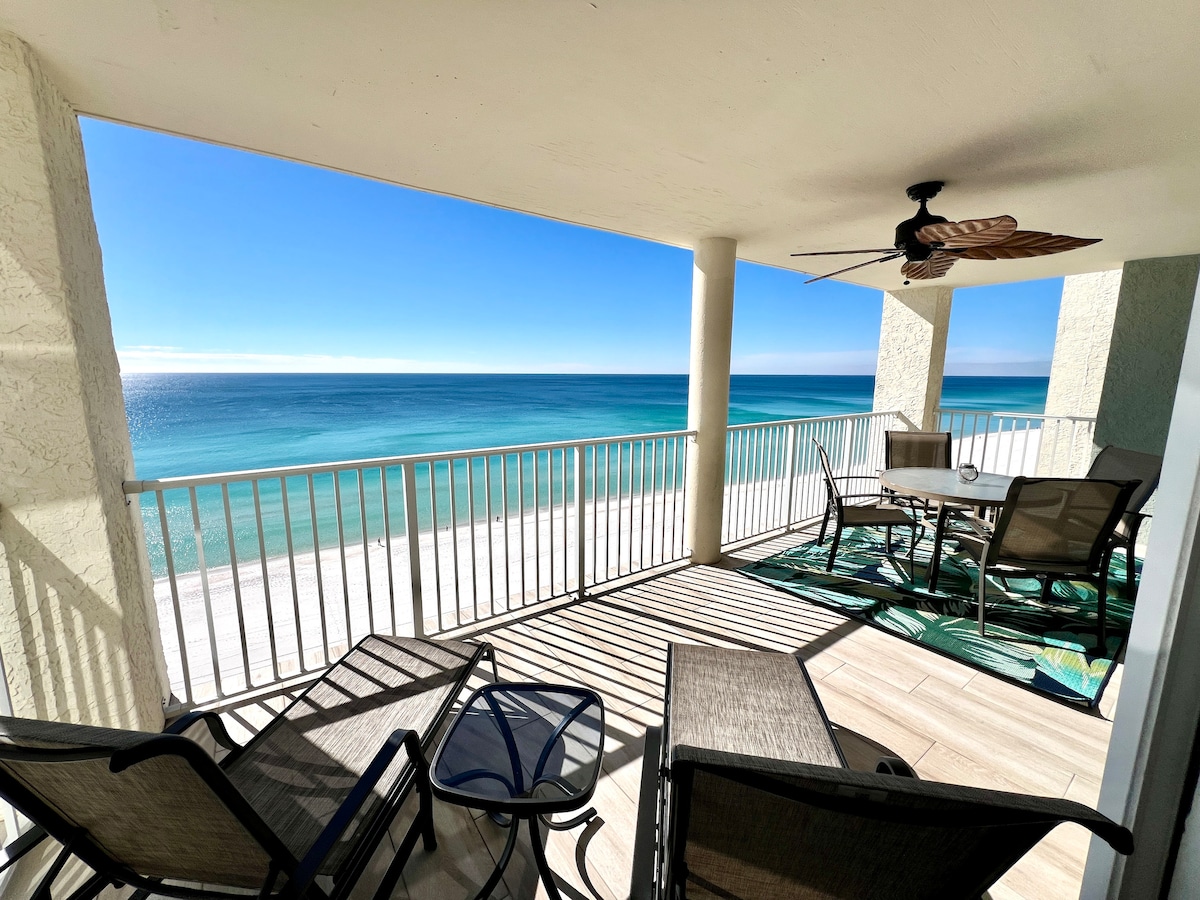 A large deck offers panoramic views of the Gulf waters, framed by columns and a ceiling fan. Seating includes comfortable chairs and a glass table, allowing for relaxation while enjoying the serene coastal scenery. Sunlight casts gentle shadows along the decking.