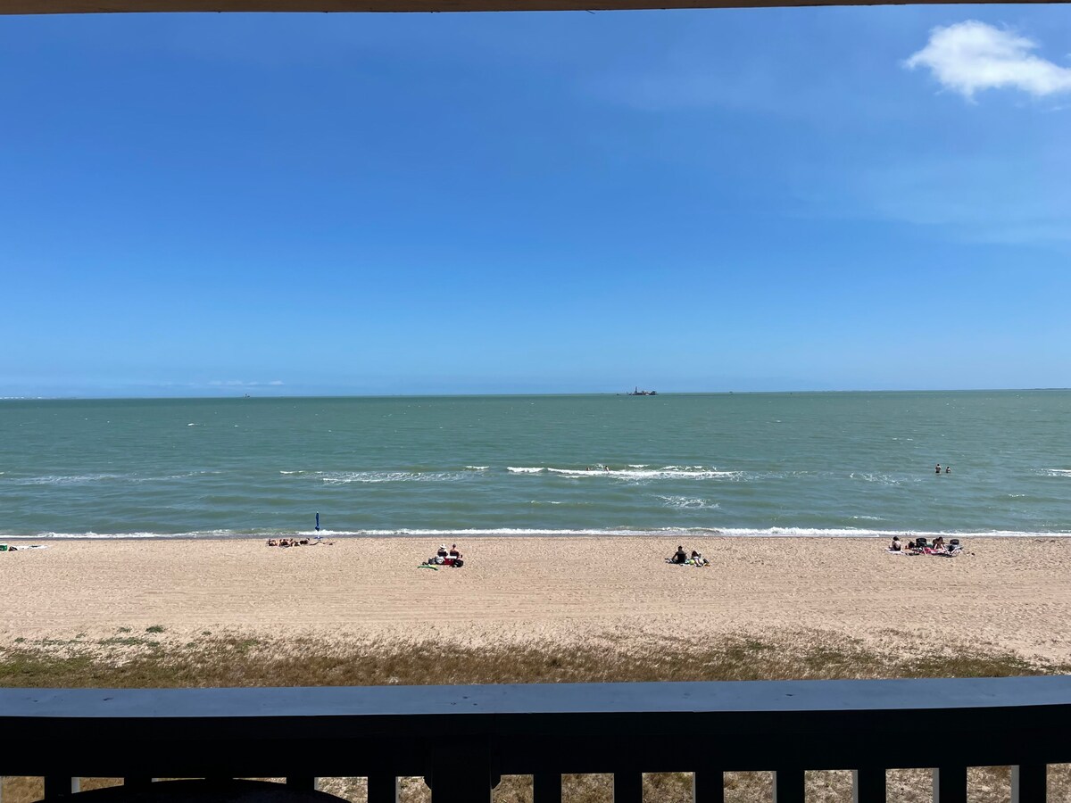 A view of the sandy beach and gentle waves under a clear blue sky is presented. People can be seen enjoying the shoreline, while a distant ship is visible on the horizon. The balcony railing provides a defined edge to this picturesque coastal scene.