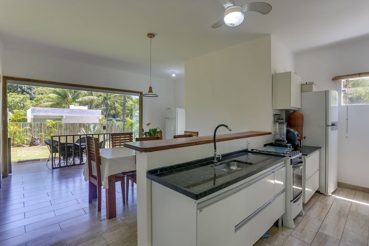 A modern kitchen is shown with sleek white cabinetry and a dark countertop. Natural light enters through a window above the sink. An adjoining dining area features a wooden table and chairs, with a view of the lush greenery outside.