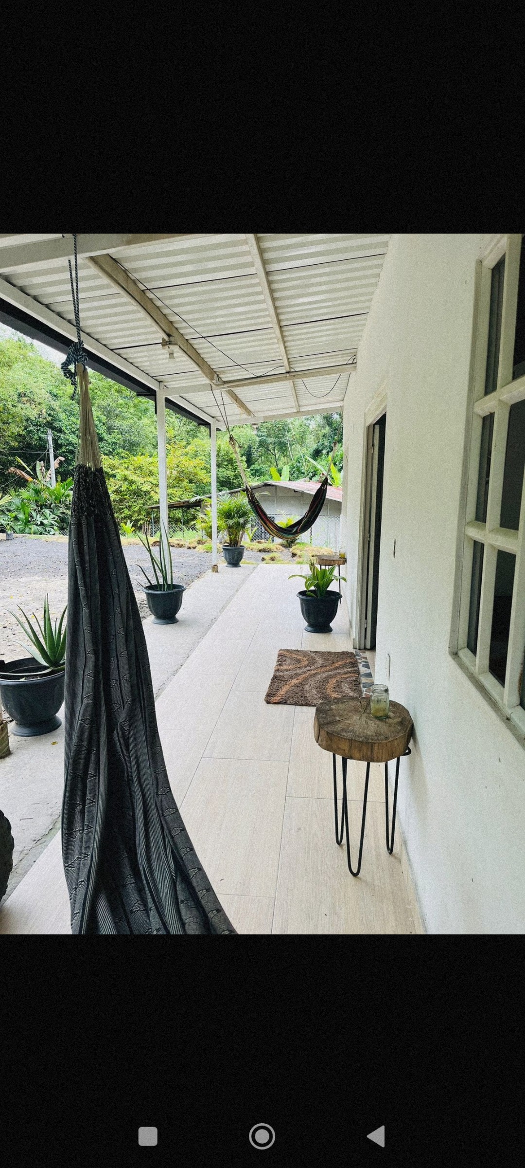A shaded terrace is framed by potted plants, creating a serene atmosphere. A hammock hangs on one side, while a small table is positioned nearby. The pathway is lined with light tiles, leading toward a natural green backdrop.