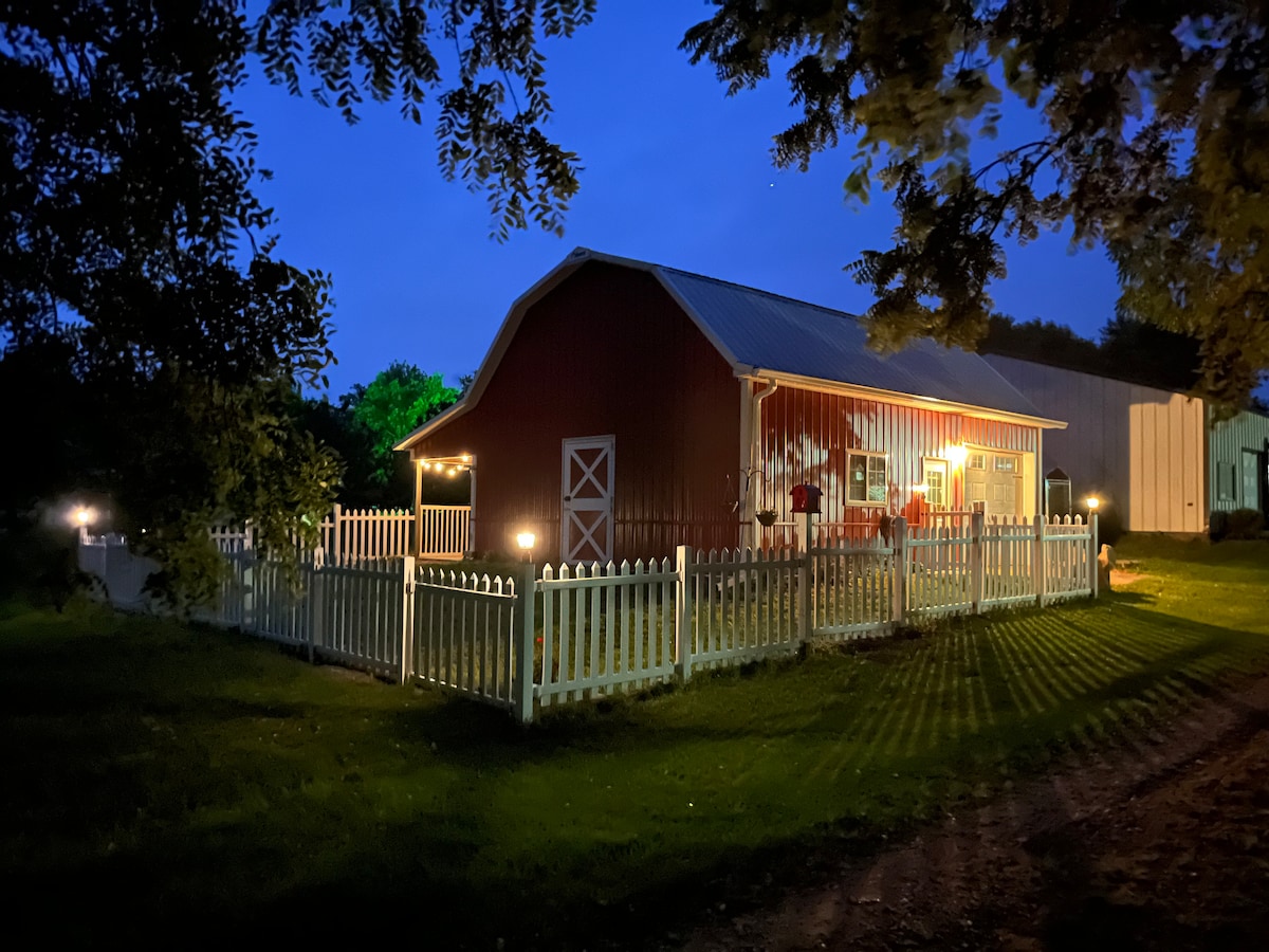 A charming red barn, illuminated by soft lights, is surrounded by a white picket fence. The evening sky displays deep blues, creating a peaceful backdrop. Shadows from the trees and fence create patterns on the grassy area, inviting guests to explore the surroundings.