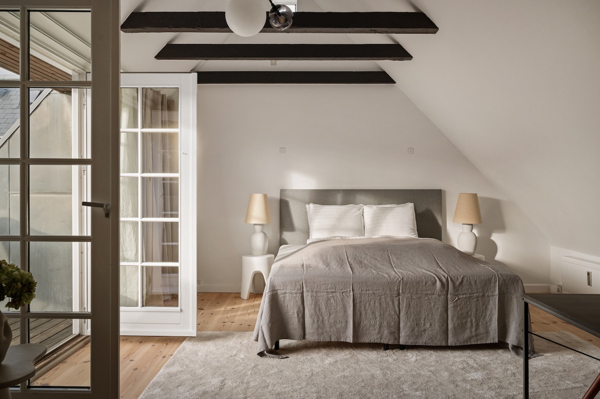 The master bedroom features a spacious bed with neutral bedding and a soft rug underfoot. Two bedside lamps provide gentle lighting, while large windows with French doors lead to a private balcony. Exposed beams add character to the ceiling.