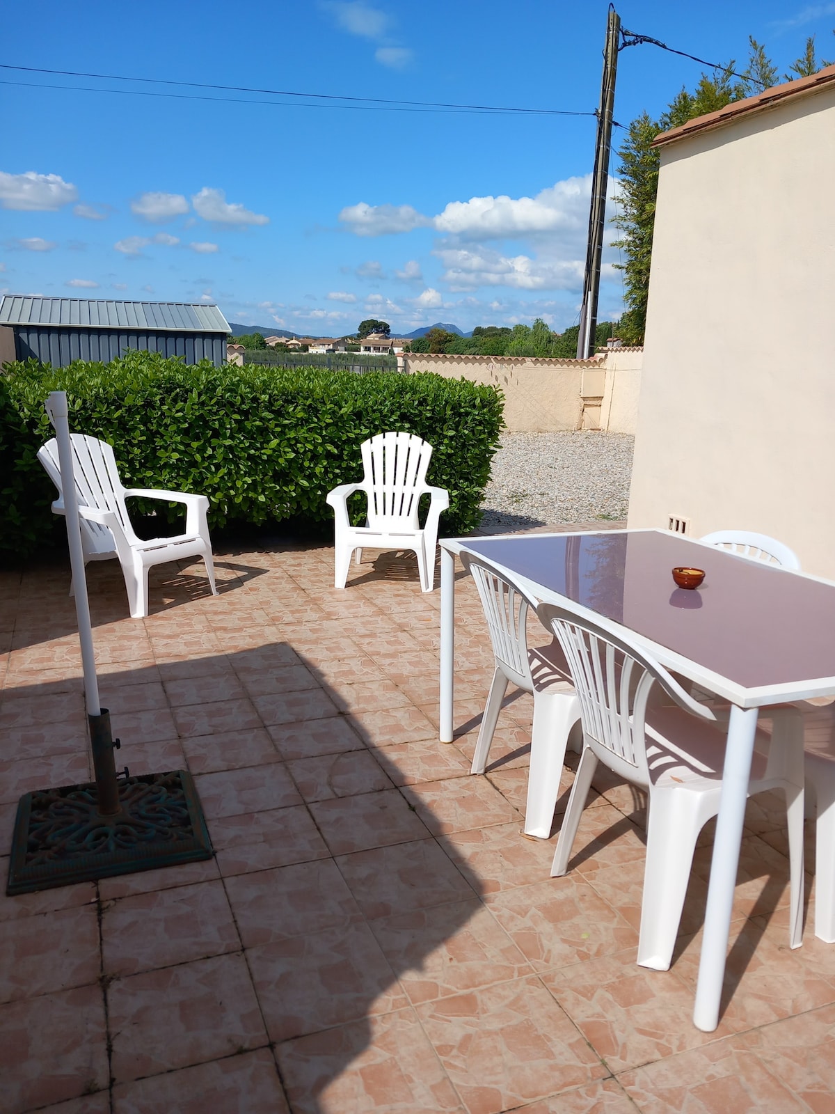 An outdoor terrace featuring a white table and four matching chairs is highlighted. A small decorative item rests on the table. Green shrubbery provides a natural backdrop, while a gravel area is visible beyond the terrace. Clear blue skies with scattered clouds can be seen.
