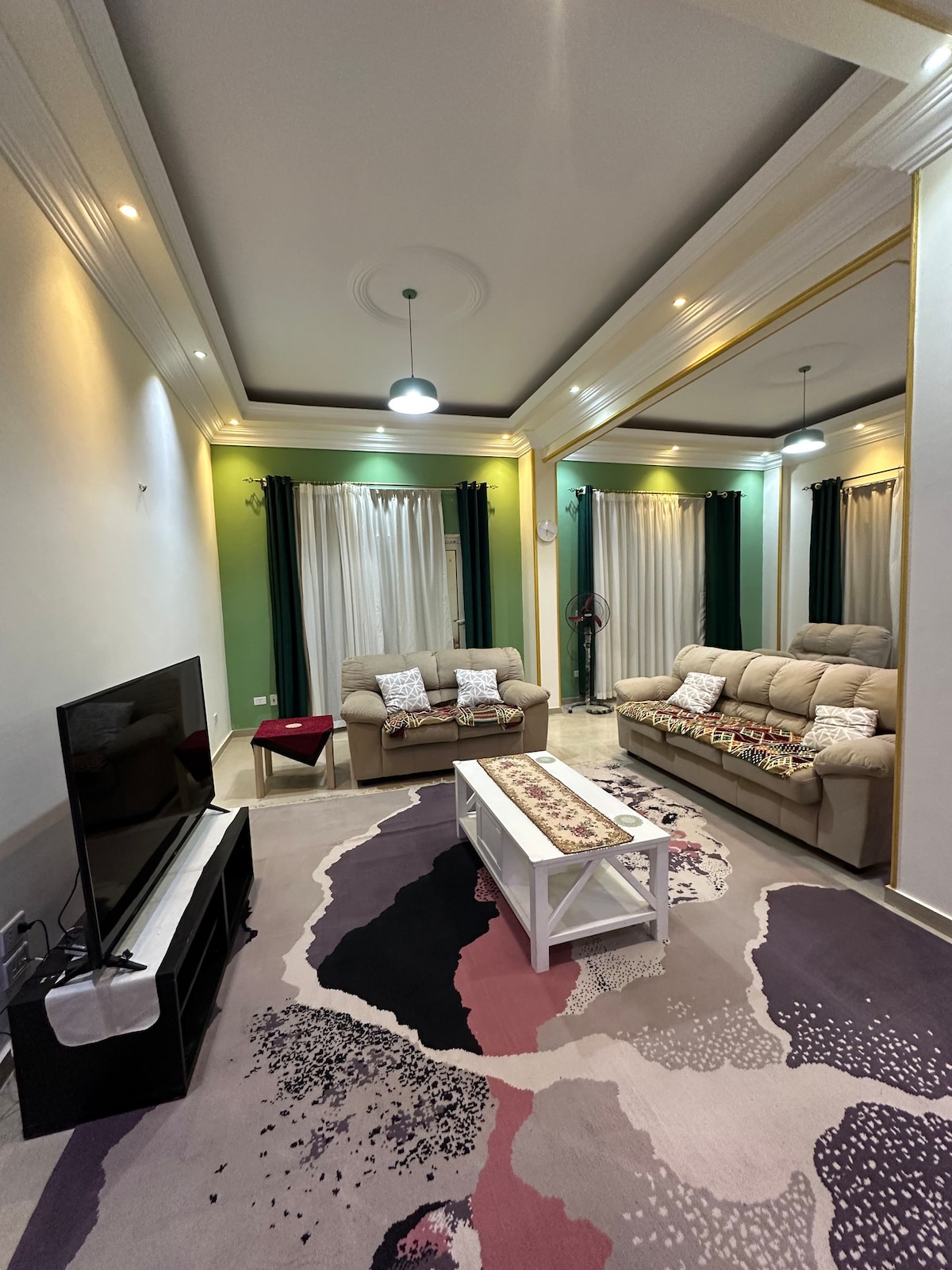 A cozy living area features a light-colored sofa set with patterned pillows and a coffee table at the center. A large television is positioned against a wall, with floor-to-ceiling curtains framing the windows, complemented by vibrant green accents in the decor.