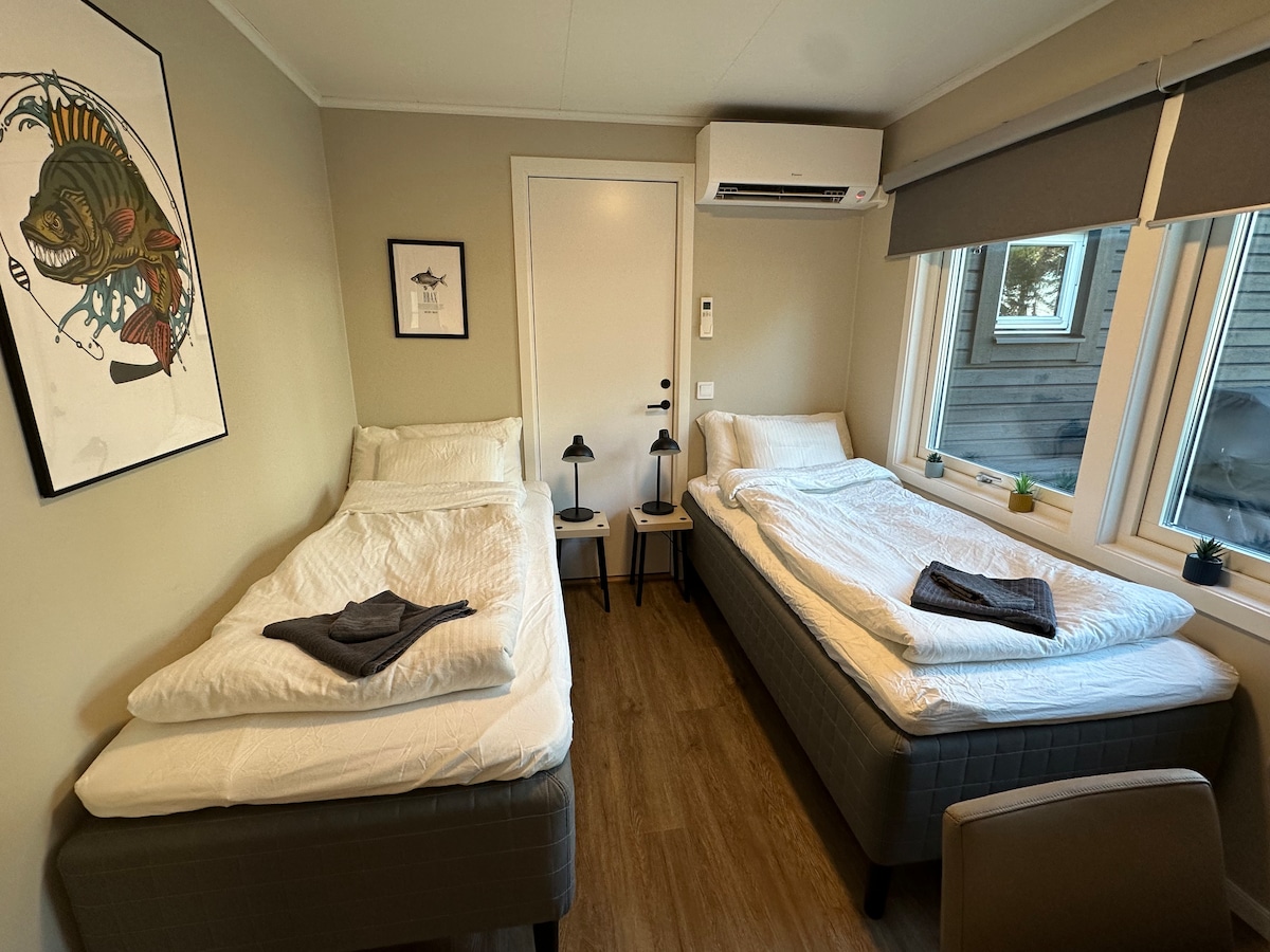 The room features two single beds neatly made with crisp white linens. Each bed is accompanied by a nightstand with a lamp. Natural light fills the space through two windows, and a wall-mounted air conditioner is installed for comfort.