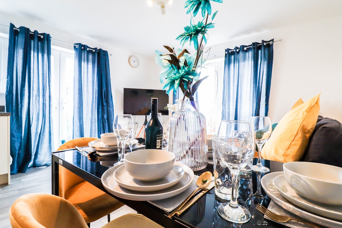 A dining area is set with elegant tableware, including plates, glasses, and cutlery. A vase with artificial flowers sits as a centerpiece. Light flows through partially drawn curtains, and a smart TV is visible in the background against a neutrally painted wall.