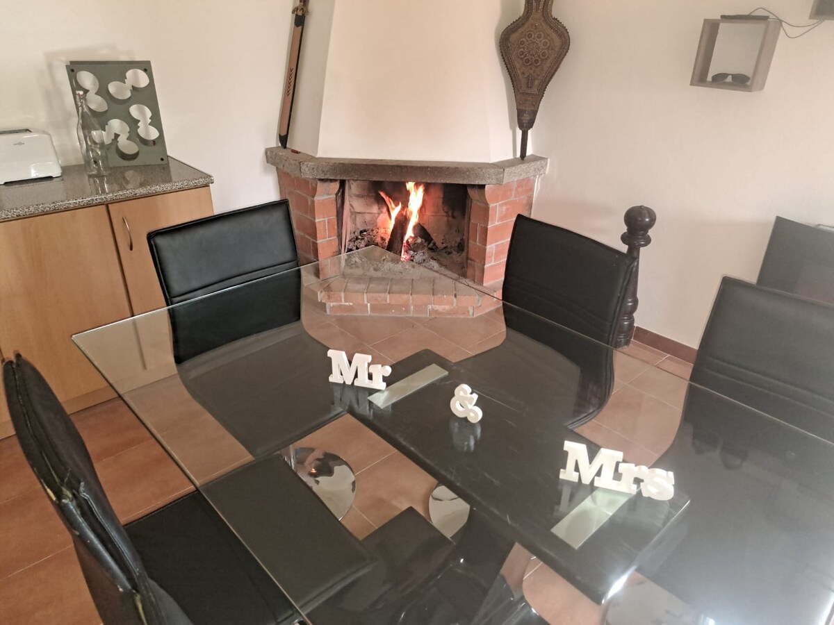 A contemporary dining area features a glass table set for six, with black leather chairs surrounding it. A traditional fireplace is visible in the background, with a warm flame flickering inside. Decorative elements include white letters spelling 'Mr & Mrs' at the table's center.