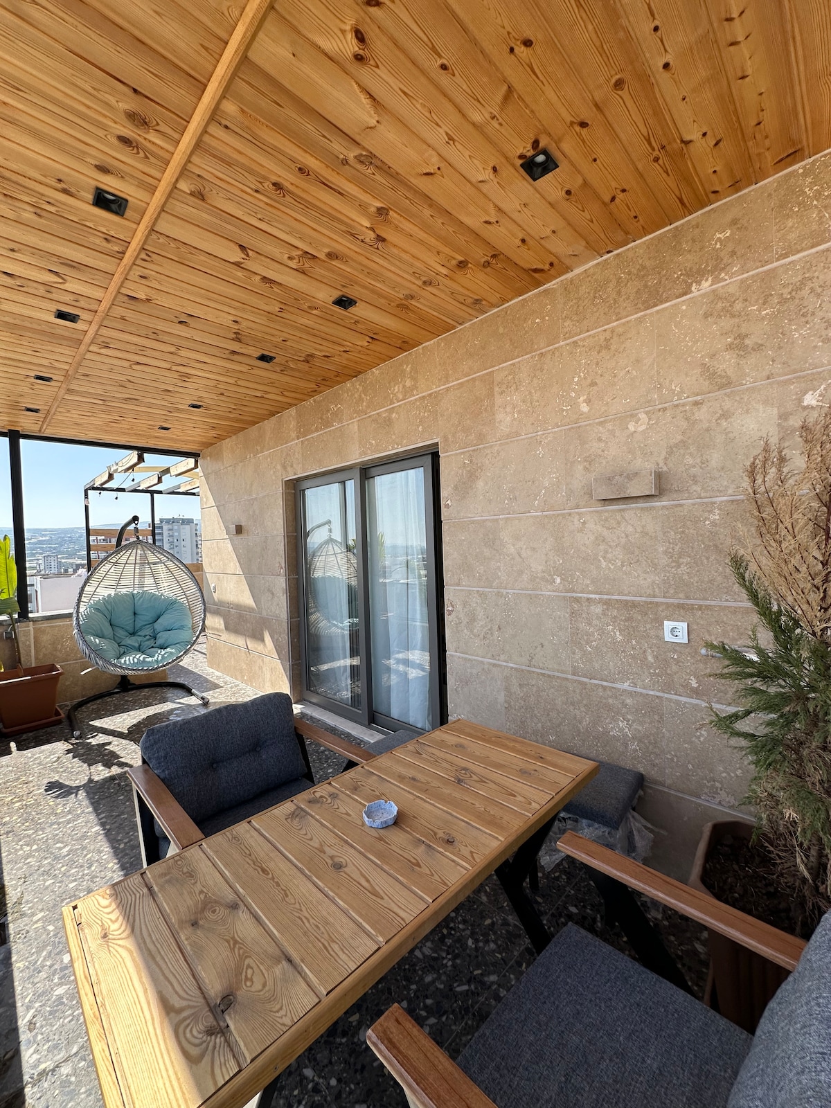 A spacious outdoor terrace features a wooden dining table complemented by two comfortable chairs. A hanging chair adds an inviting touch, while the ceiling showcases natural wood panels. Potted plants enhance the serene atmosphere, with city views visible through the glass doors.