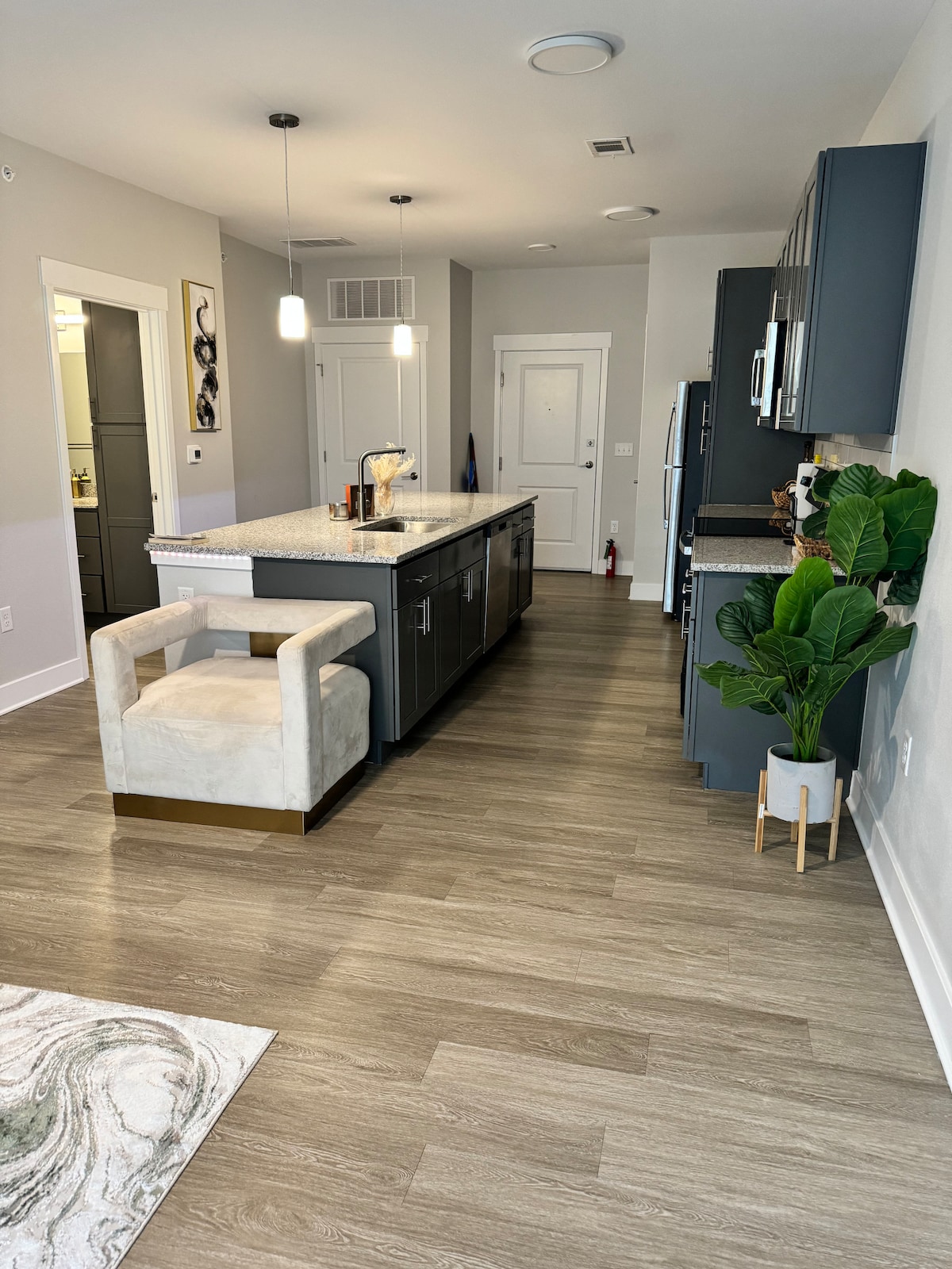 A modern kitchen is equipped with a central island topped with light-colored granite. Adjacent cabinets are finished in a deep hue. A plush chair sits beside the island, and a small potted plant adds a touch of greenery. The space is well-lit with pendant lighting.