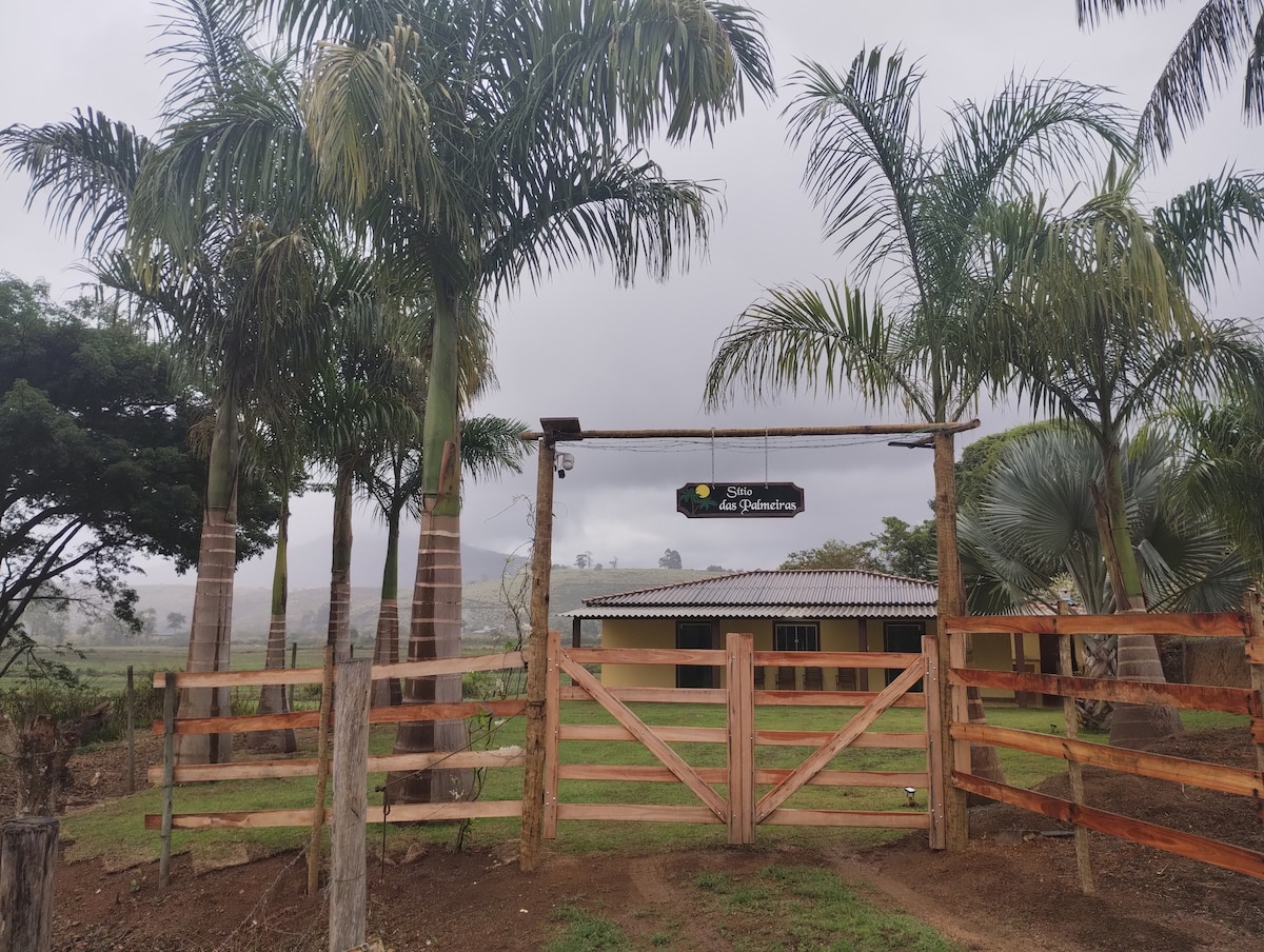 A wooden gate opens to a spacious outdoor area, framed by tall palm trees. The structure features a covered area with a roof that complements the natural landscape. A sign indicating the property name is prominently displayed above the entrance.