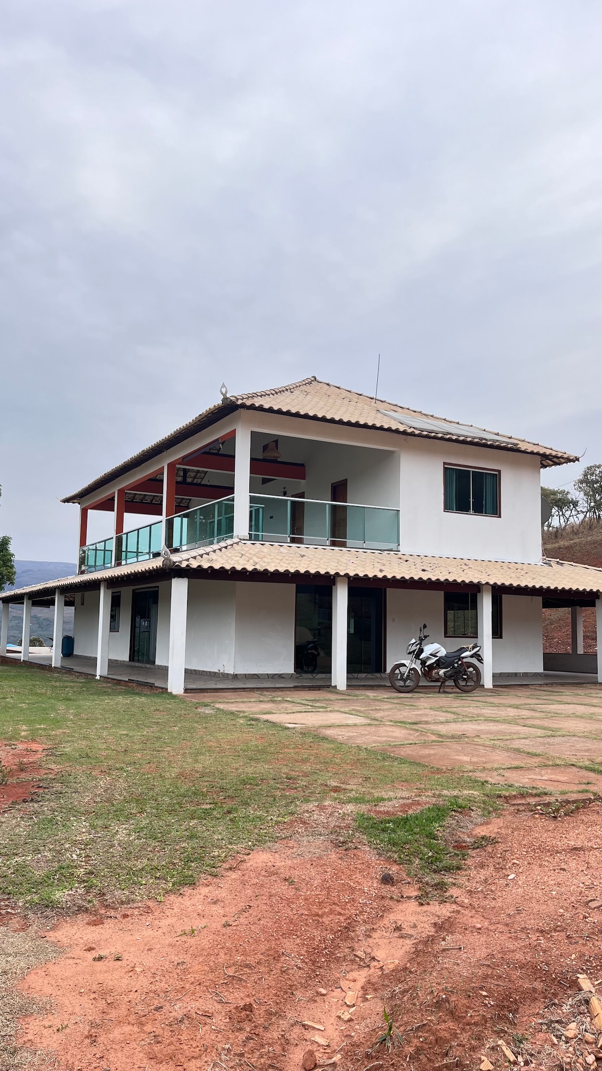 A two-story house is presented, featuring a white exterior with a tiled roof. Large glass doors provide access to a spacious balcony on the second floor. A motorcycle is parked near the entrance, while a grassy area surrounds the building.
