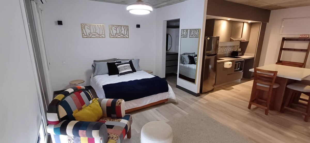 A compact living area features a spacious bed draped with a navy blue throw and accented by decorative pillows. A colorful, patterned armchair adds a pop of color. The kitchenette and dining area are visible in the background, characterized by modern appliances and natural wood finishes.