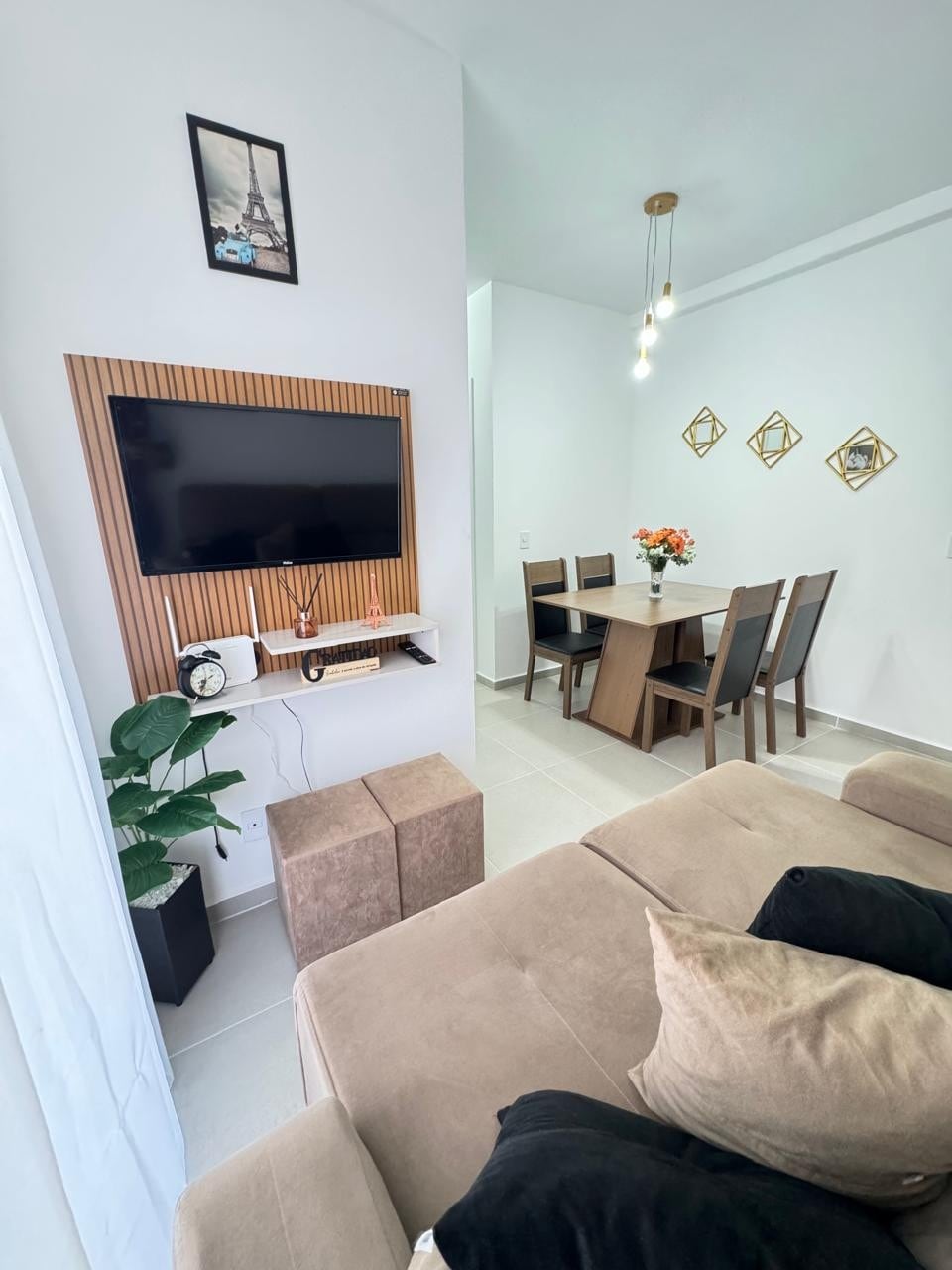 A modern living area features a mounted television, framed art, and a cozy beige sofa with decorative pillows. A dining table with six chairs sits nearby, complemented by a floral centerpiece. Natural light illuminates the space, accentuating the contemporary decor.