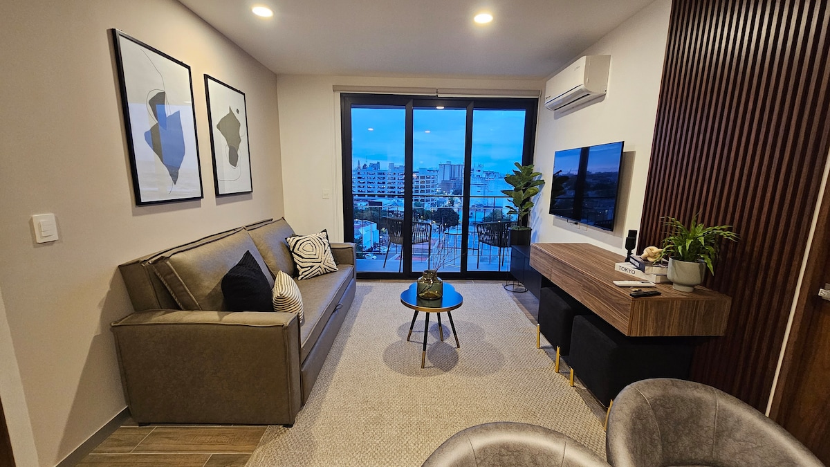 A modern living area features a comfortable sofa paired with decorative cushions. A minimalist coffee table sits in front, complemented by a stylish plant. Large glass doors open to a balcony, providing a view of the city. A wall-mounted television and artwork enhance the ambiance.