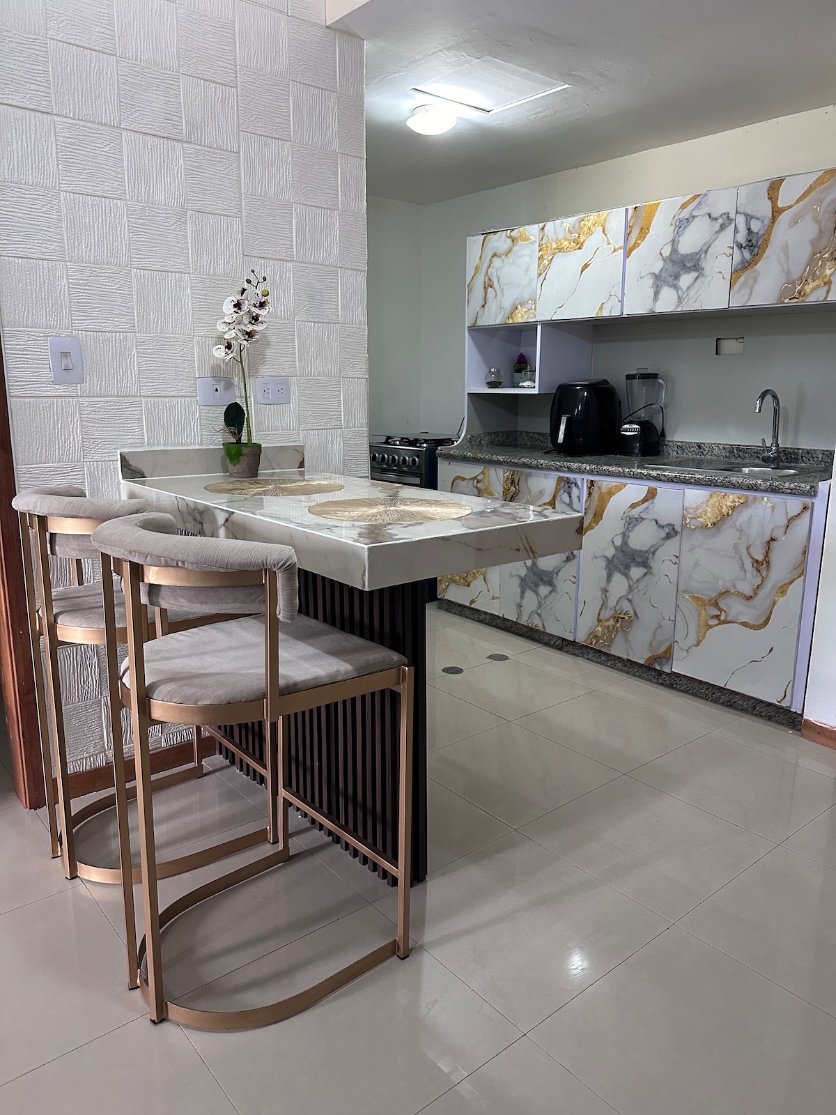 A modern kitchen space is highlighted by elegant marble-patterned cabinetry. A small island with two stools provides seating. A sleek black coffee maker and a stove are visible, alongside a spacious sink and countertop, all complemented by soft lighting.