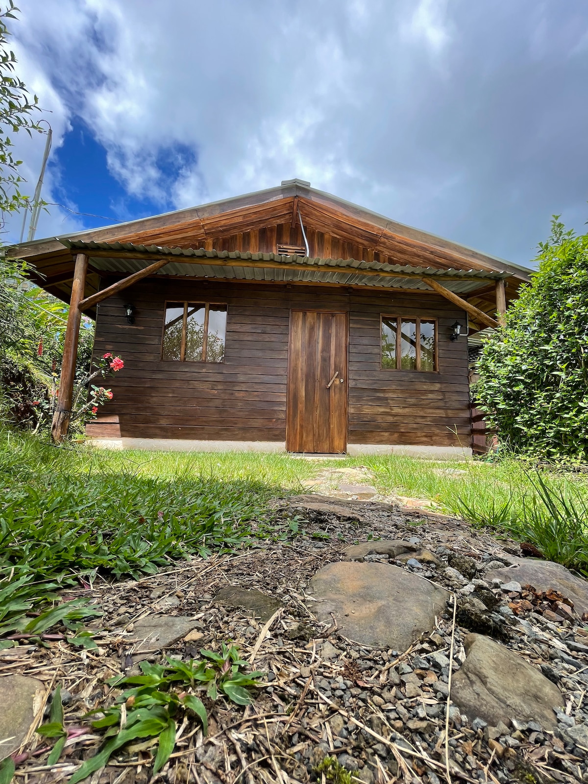 A charming wooden cabin is set within a lush green landscape, showcasing a balcony and two large windows. The door is made of wood and blends seamlessly with the cabin's natural aesthetics, while a stone pathway leads to the entrance.