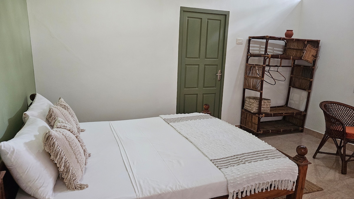 A well-appointed guest room features a spacious bed adorned with decorative pillows. A light-colored blanket complements the bed linens. A green accent wall adds a touch of calm, while a wicker chair and an open clothes rack provide convenience in the corner.