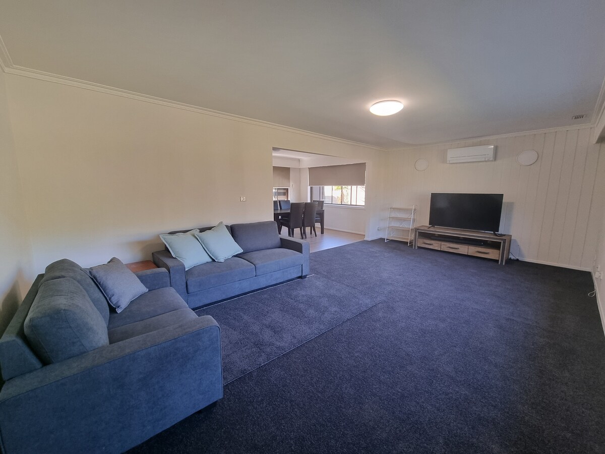 A spacious living area features a comfortable gray sectional sofa complemented by light pillows. A large flat-screen TV sits on a wooden media console. The room is carpeted in dark blue, with natural light flowing in from the adjacent dining space.