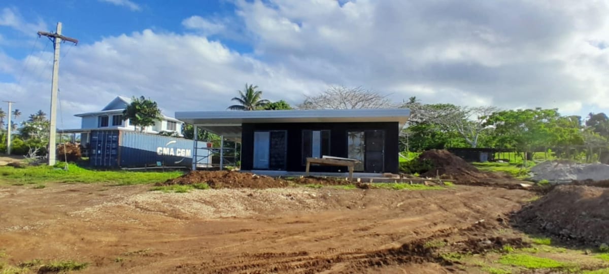 A newly constructed villa is shown with a simple, modern design. The structure features large windows and sliding doors, accented by a concrete porch. Surrounding areas are in a natural state, with soil and some greenery visible, indicating ongoing landscape work.