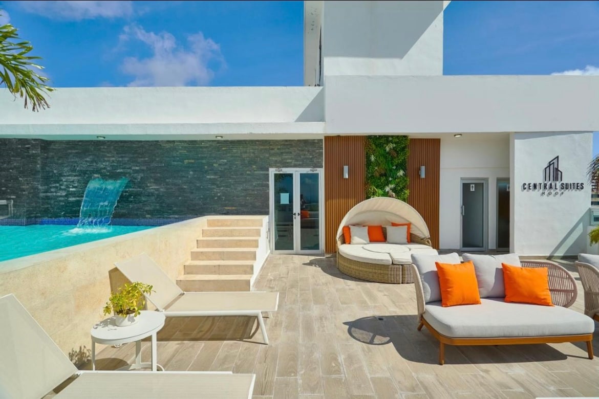 A spacious patio features modern outdoor furniture, including a circular lounge bed with bright orange cushions. Stairs lead to a serene blue pool with a calming water feature. The entrance to Central Suites is visible, surrounded by lush greenery.