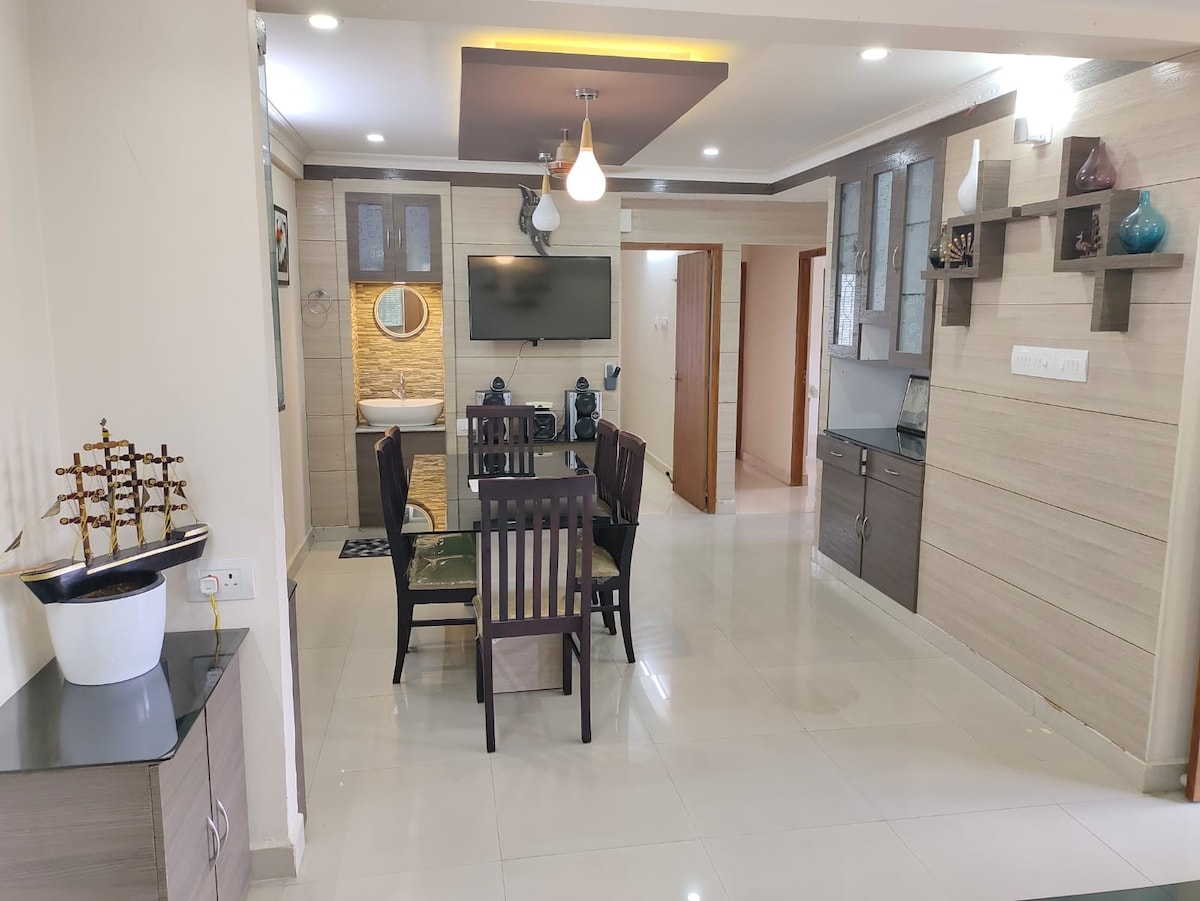 An open-plan dining area features a dark wood table surrounded by four chairs. A flat-screen television is mounted on the wall, and a small kitchenette is visible to one side. Soft lighting enhances the modern design of the space with neutral-toned walls.