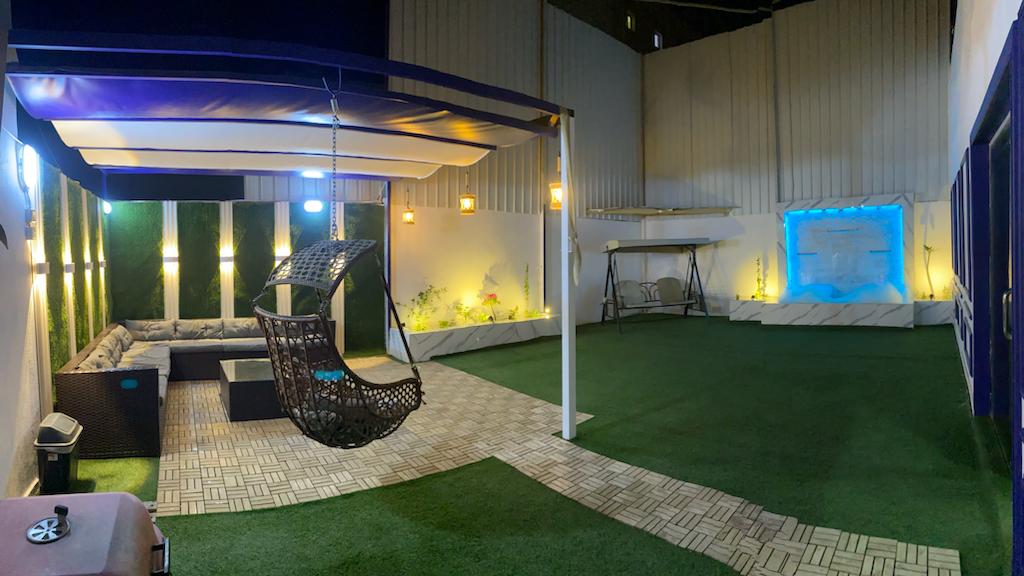 An outdoor space is highlighted by a hanging chair and a comfortable sectional sofa surrounding a central coffee table. Soft lighting creates an inviting atmosphere, while greenery complements the decorative features. A well-maintained artificial lawn adds to the relaxing environment.