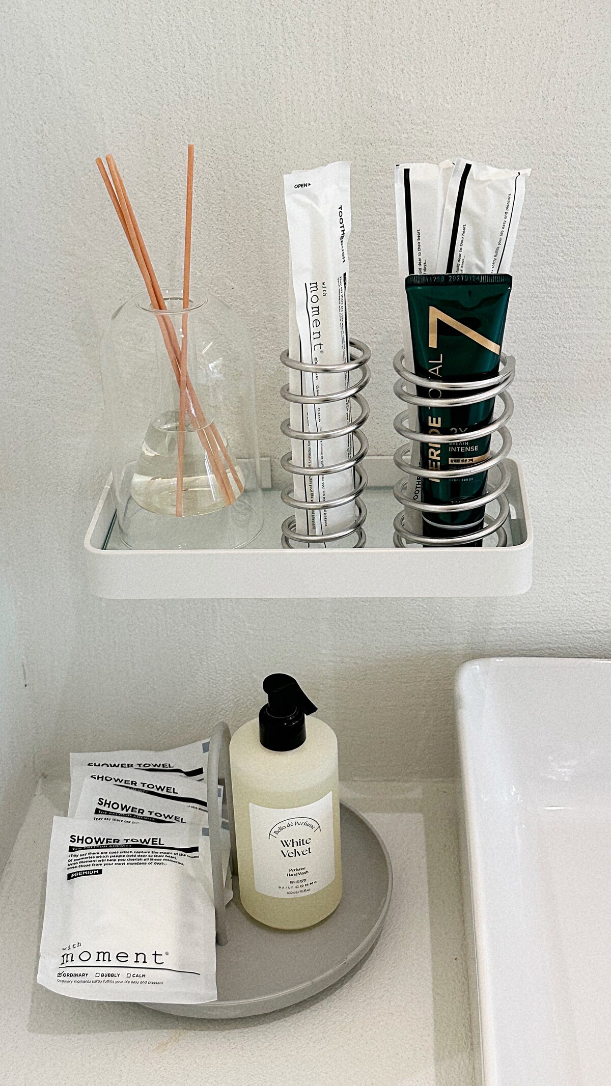 A minimalist bathroom shelf is displayed, featuring a clear glass vase with decorative sticks, standing alongside two holders containing toothpaste and a tube of skincare product. A circular tray holds neatly packaged toiletries, while a pump bottle is positioned nearby.