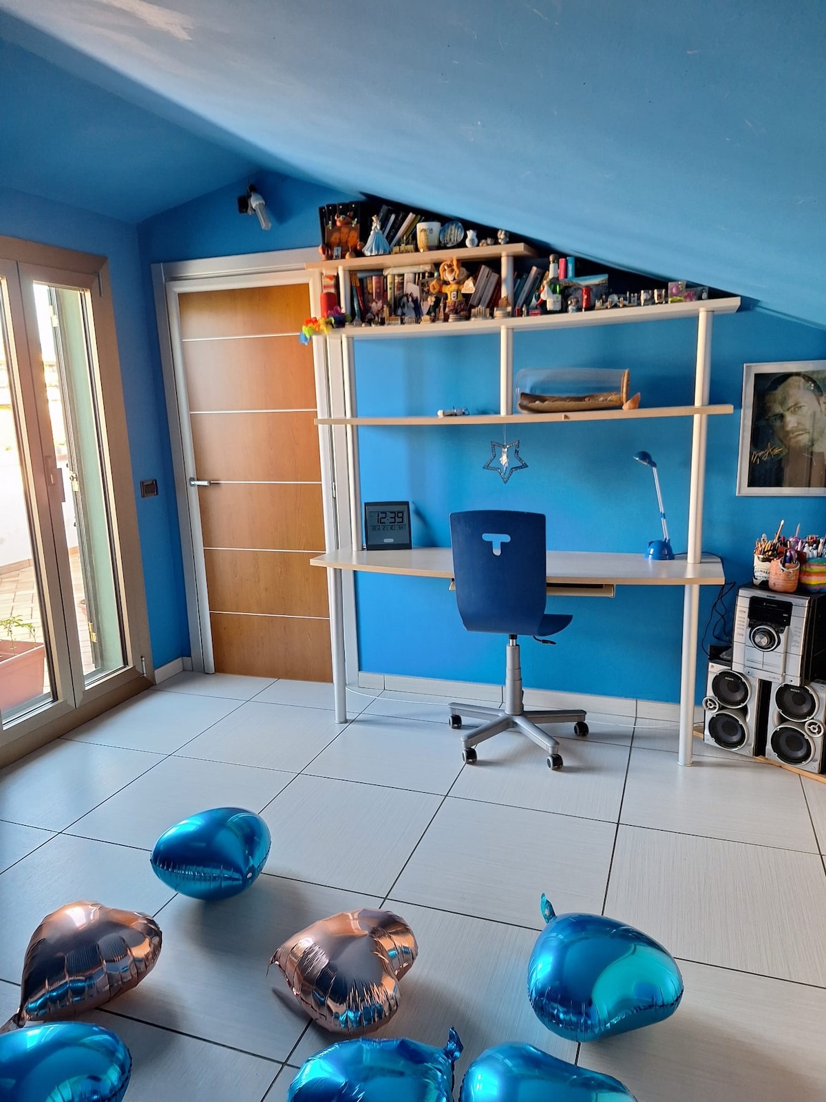 A bright blue room is featured, with a desk and chair positioned neatly against the wall. Shelves above the desk hold various items. Large windows allow natural light, and metallic blue balloons are scattered on the floor, creating a festive atmosphere.