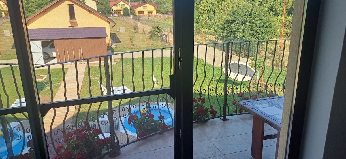 A balcony view is presented, featuring a green lawn with lounge areas and a swimming pool visible below. Decorative flowers are seen along the balcony railing, complementing the outdoor space. A table and chairs are positioned for seating, enhancing the area for relaxation.