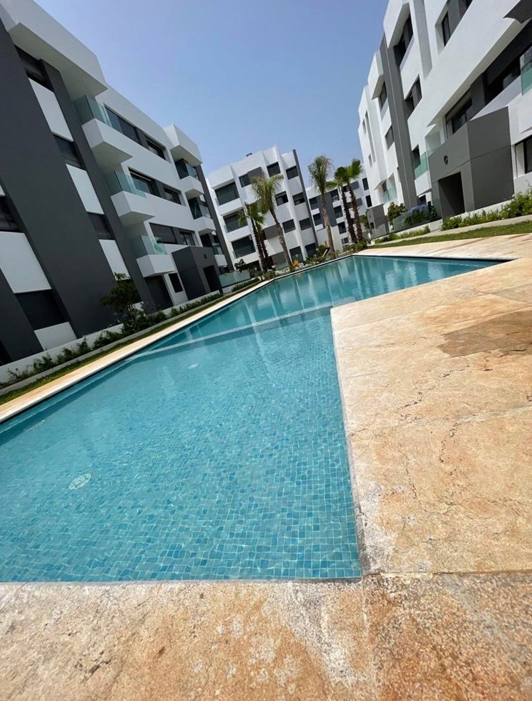 A serene swimming pool is framed by modern, white and grey buildings. The pool's clear water reflects the sky, while landscaped greenery surrounds the area, enhancing the tranquil setting. Sun loungers are positioned along the poolside, inviting relaxation and enjoyment.