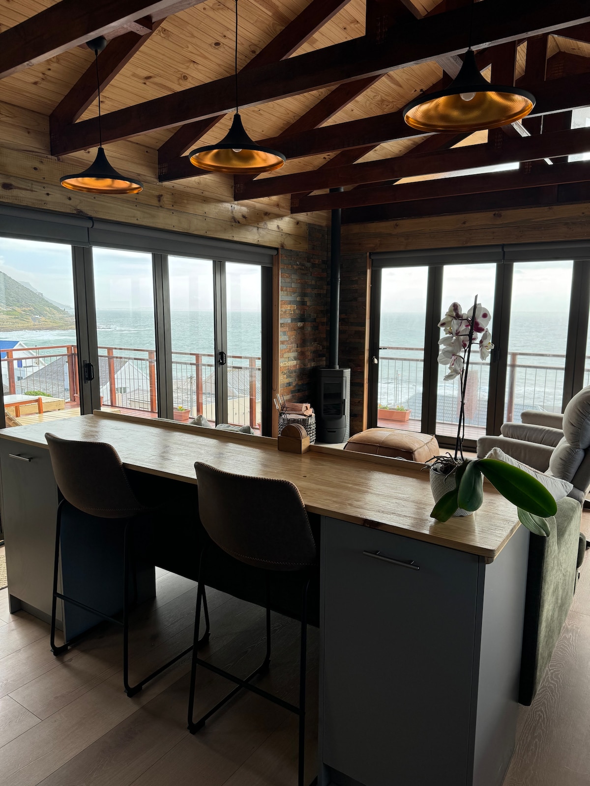 A bright interior space is highlighted by wooden ceiling beams and modern pendant lighting. A counter with seating for two offers a view through large glass doors leading to an outdoor area overlooking the water. Subtle greenery adds a natural touch to the scene.