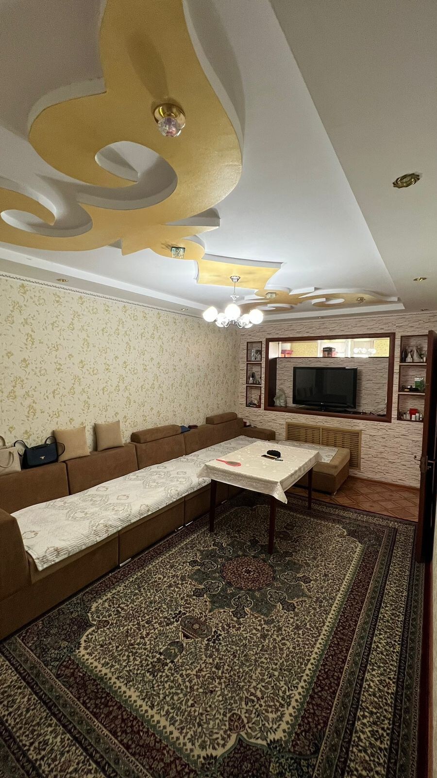 A spacious living area features a central table surrounded by a light-colored sofa set. Decorative wall accents and a patterned area rug enhance the room's character. A chandelier hangs from the ceiling, providing ambient lighting, while a built-in shelf displays decorative items and a television.