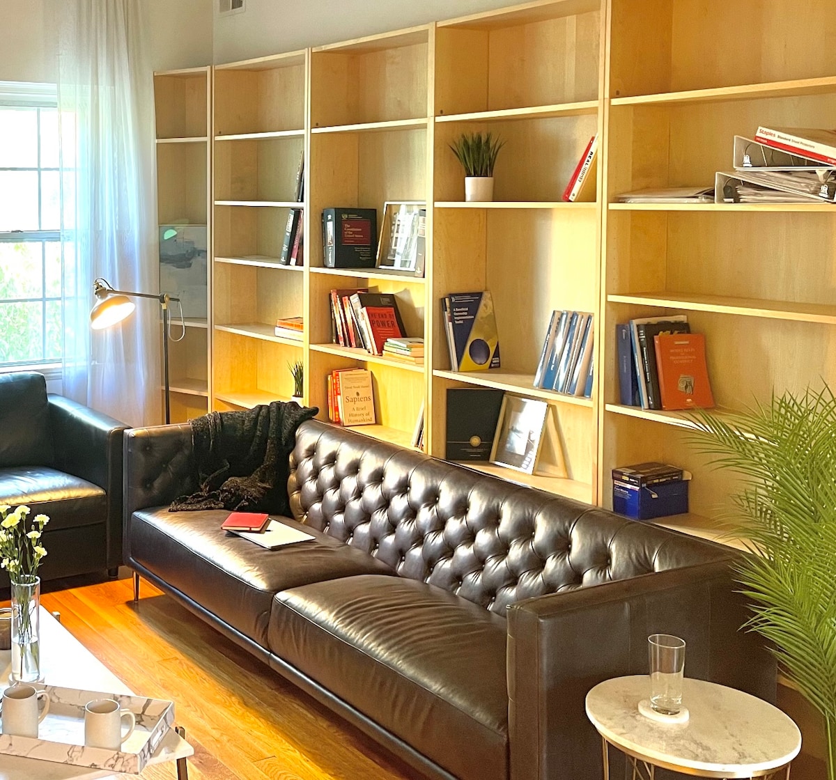 A cozy living area features a dark leather sofa facing a wooden bookshelf filled with a variety of books and decorative items. A side table holds a glass and a small plant, while a floor lamp provides warm lighting. Natural light enters through the window.