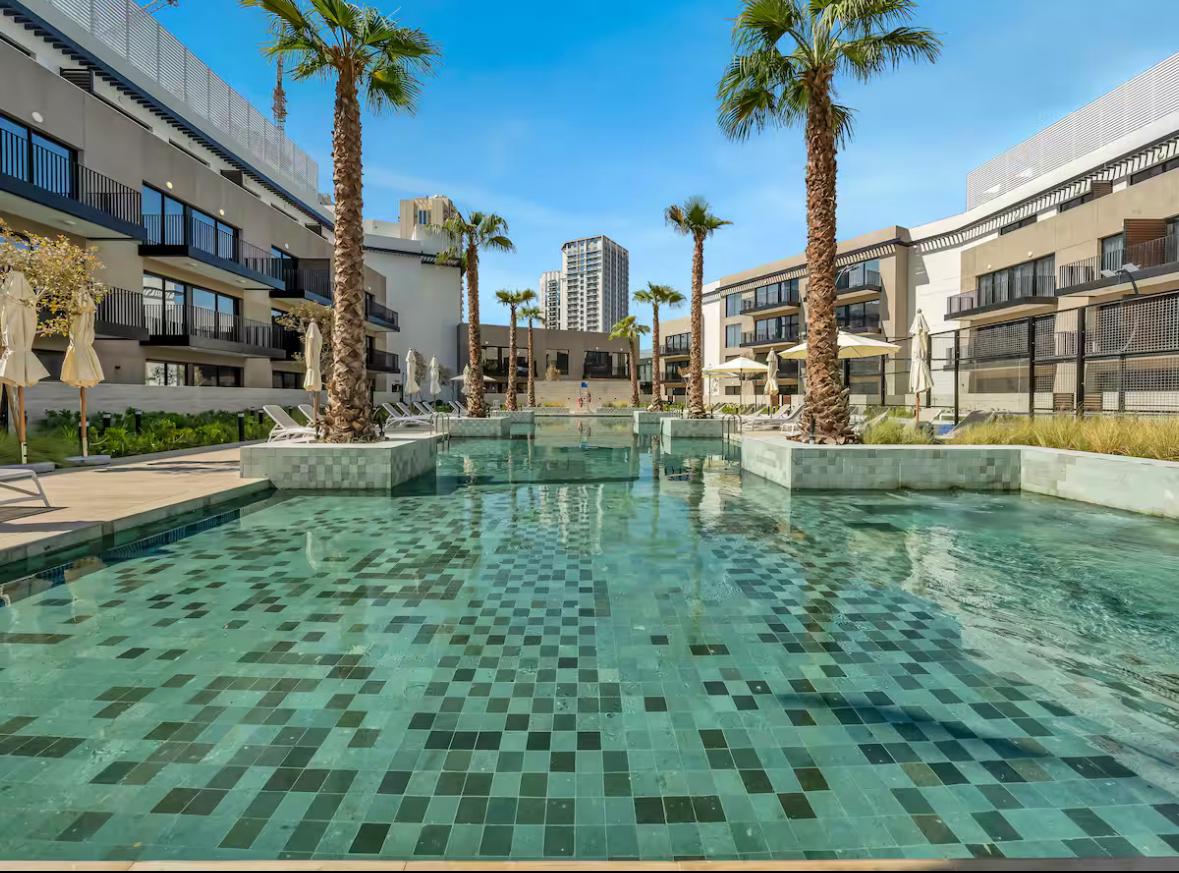 A tranquil pool area features palm trees and modern loungers, surrounded by stylish buildings. The water's surface reflects tiles in varying shades of green, creating a calming visual. Sunlight illuminates the space, enhancing the inviting atmosphere of relaxation and leisure.