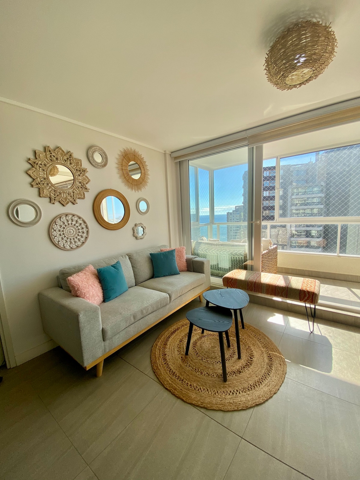 A light-filled living area features large windows with ocean views. A gray sofa adorned with teal and peach cushions is positioned beside a small round table. Wall-mounted mirrors in various sizes add visual interest, while a woven rug grounds the seating space.