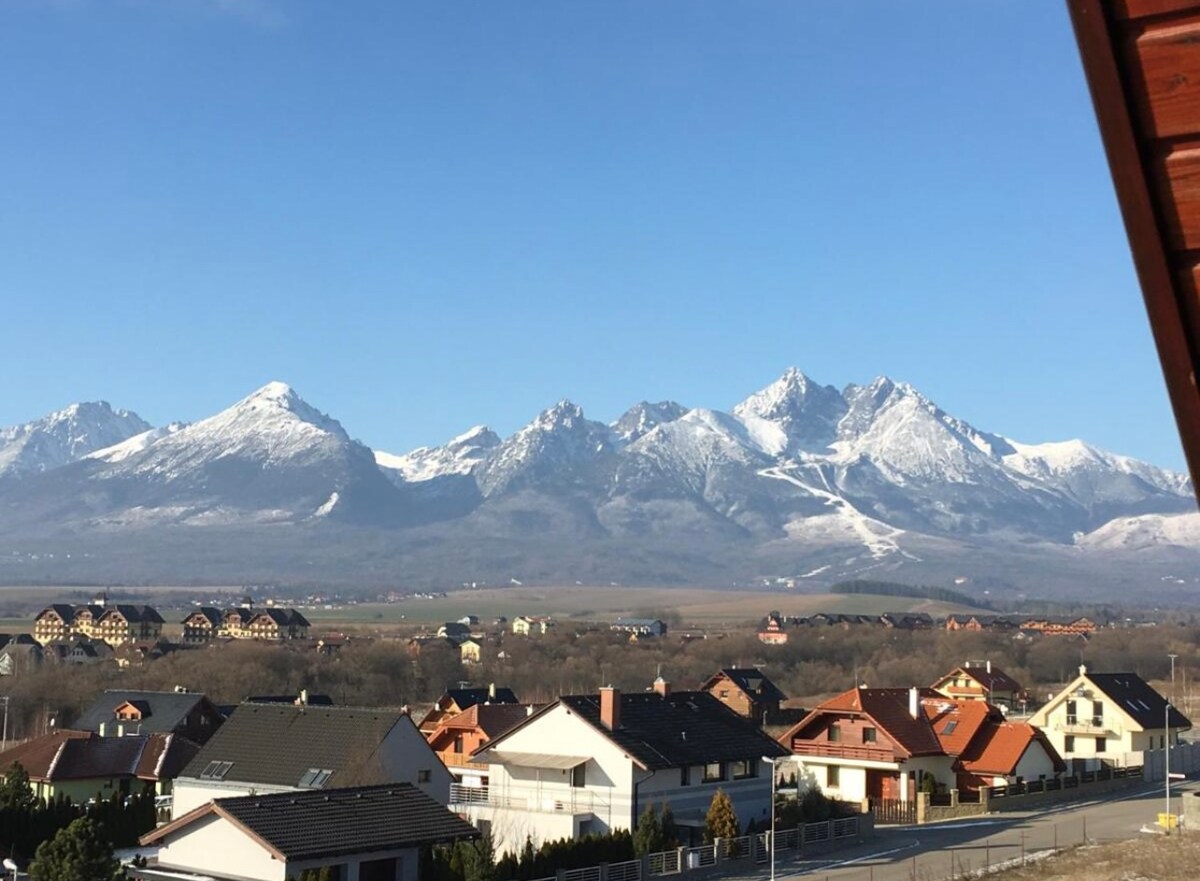 A scenic view of the Tatry mountains is presented in the background, featuring snow-capped peaks under a clear blue sky. In the foreground, a collection of houses with varied architectural styles is visible, set against the natural landscape.