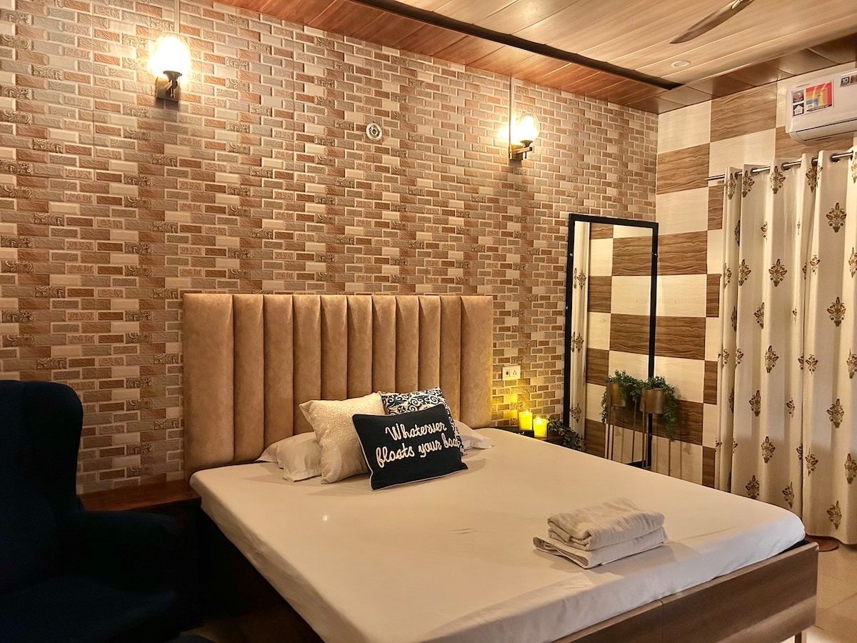 The bedroom features a spacious bed with a plush headboard and neutral bedding. A cozy armchair is positioned nearby. Decorative wall lighting and textured brick wallpaper contribute to the room's calming atmosphere. A mirror reflects the room, enhancing the sense of space.
