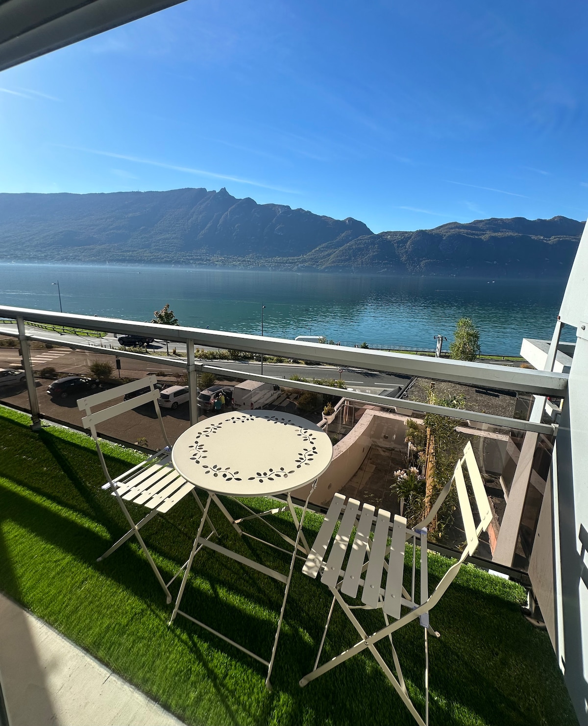 A balcony is shown, featuring a round table and two folding chairs. The vibrant green grass covering the floor complements the scenic view of the lake and mountains in the background, illuminated by clear blue skies.
