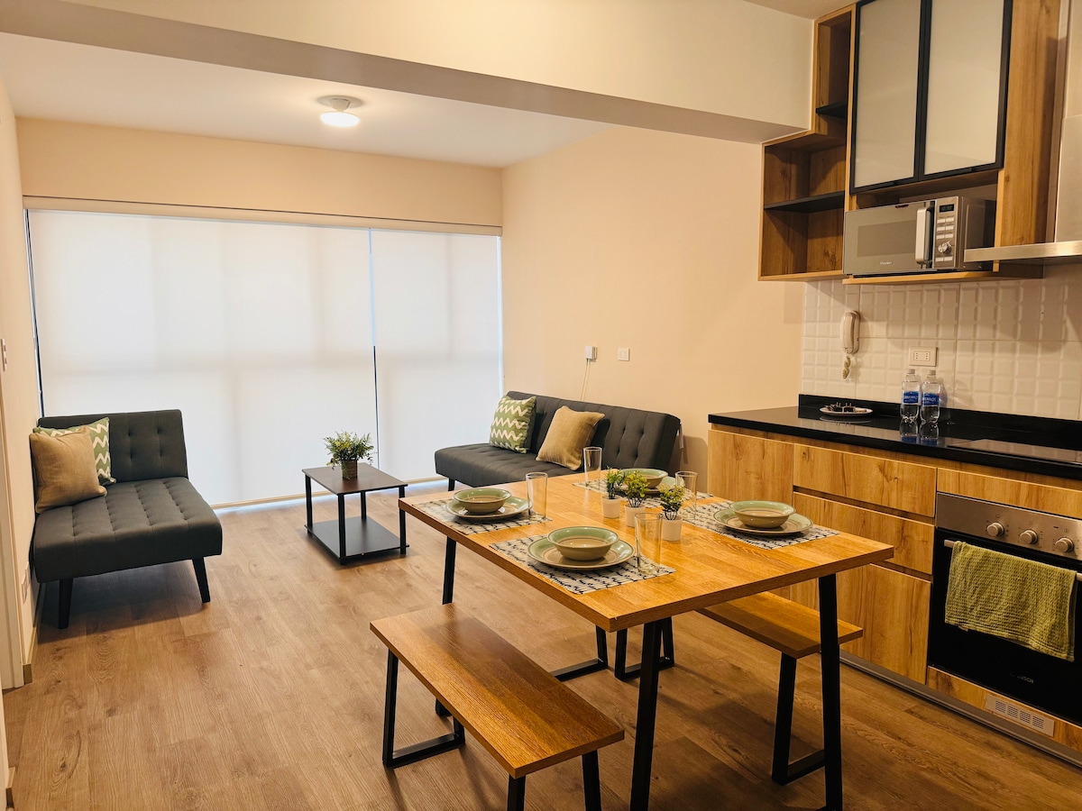 An open living and dining area is presented, featuring a wooden dining table set for four. Two stylish sofas are arranged in the seating area, complemented by subtle green accents. The kitchen space includes modern appliances, with ample cabinetry and a clean, contemporary design.