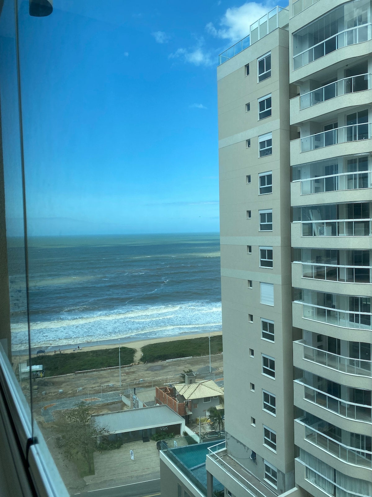 A view from a high-rise window showcases the ocean and coastal landscape. Waves gently meet the shore, while multiple buildings are visible in the foreground. The sky is bright and clear, enhancing the serene coastal atmosphere.
