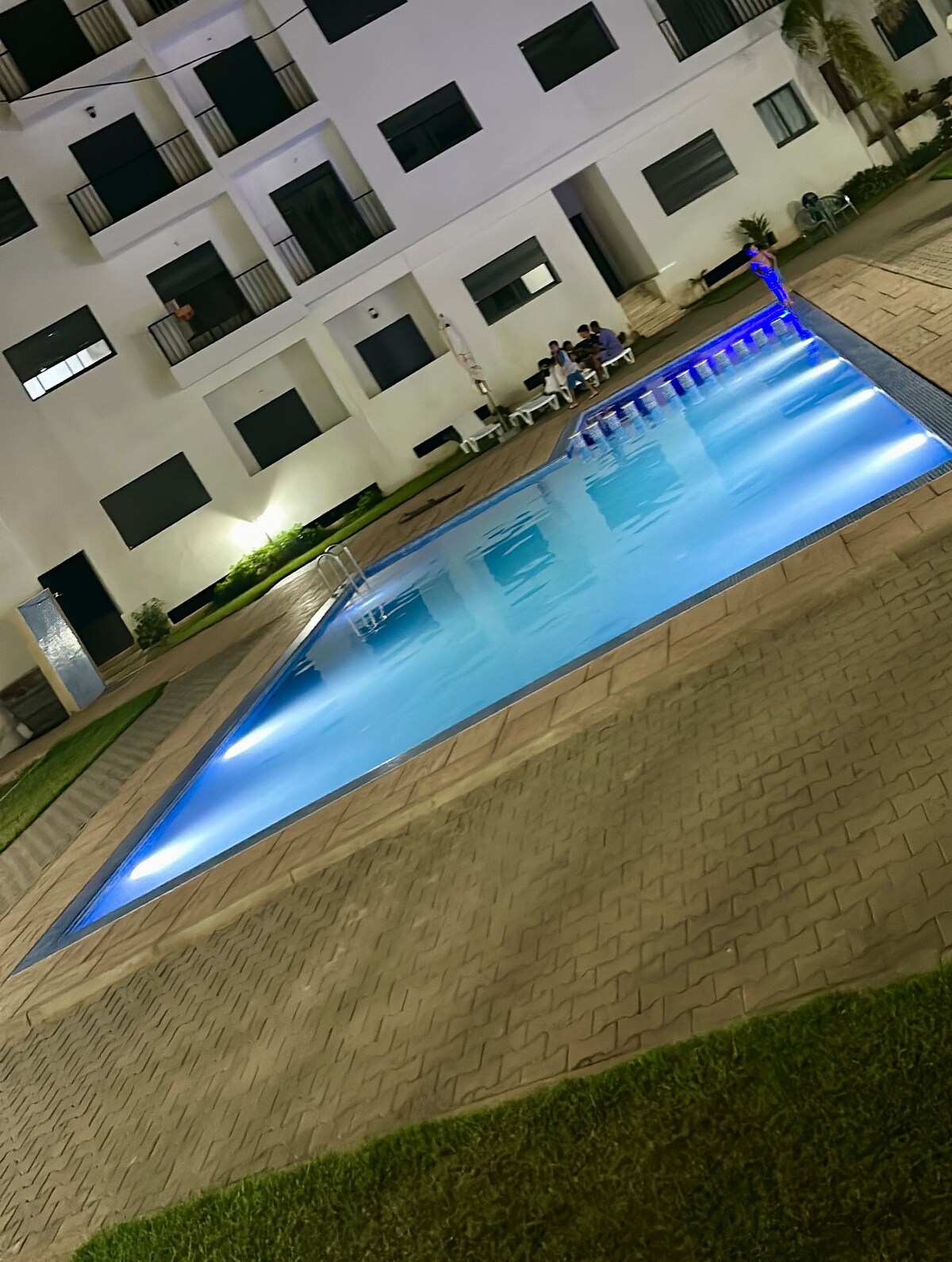 A well-lit swimming pool is surrounded by a manicured lawn and patio area. The pool features a gentle incline with steps, and is enhanced by blue LED lighting. An apartment building with several balconies provides a backdrop to the serene setting.