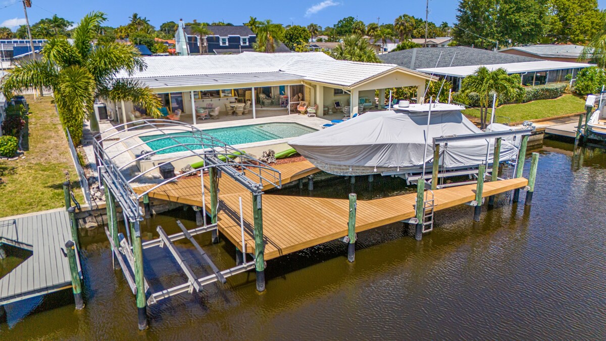 An aerial view of the home showcases the spacious outdoor area, featuring a heated pool bordered by coral stone tiles. A large dock extends into the water, equipped with a boat lift, providing ample space for fishing or launching kayaks. Lush greenery surrounds the property.
