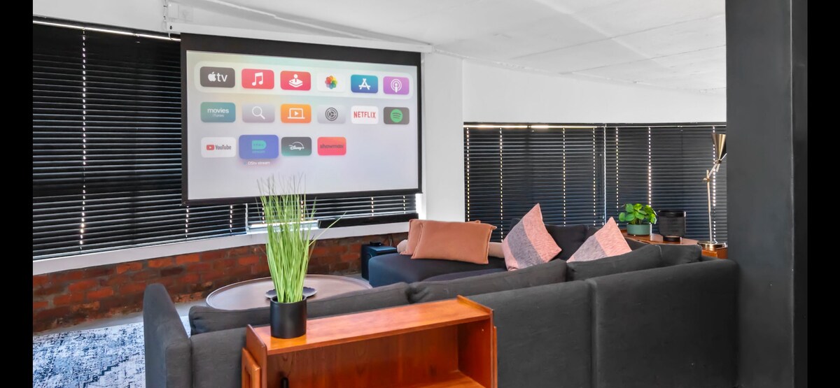 A spacious living area features a large screen displaying various entertainment options. Comfortable seating is arranged with cushions in pastel hues. Natural light is filtered through dark blinds, creating an intimate setting. A modern coffee table is surrounded by a lush plant for added greenery.
