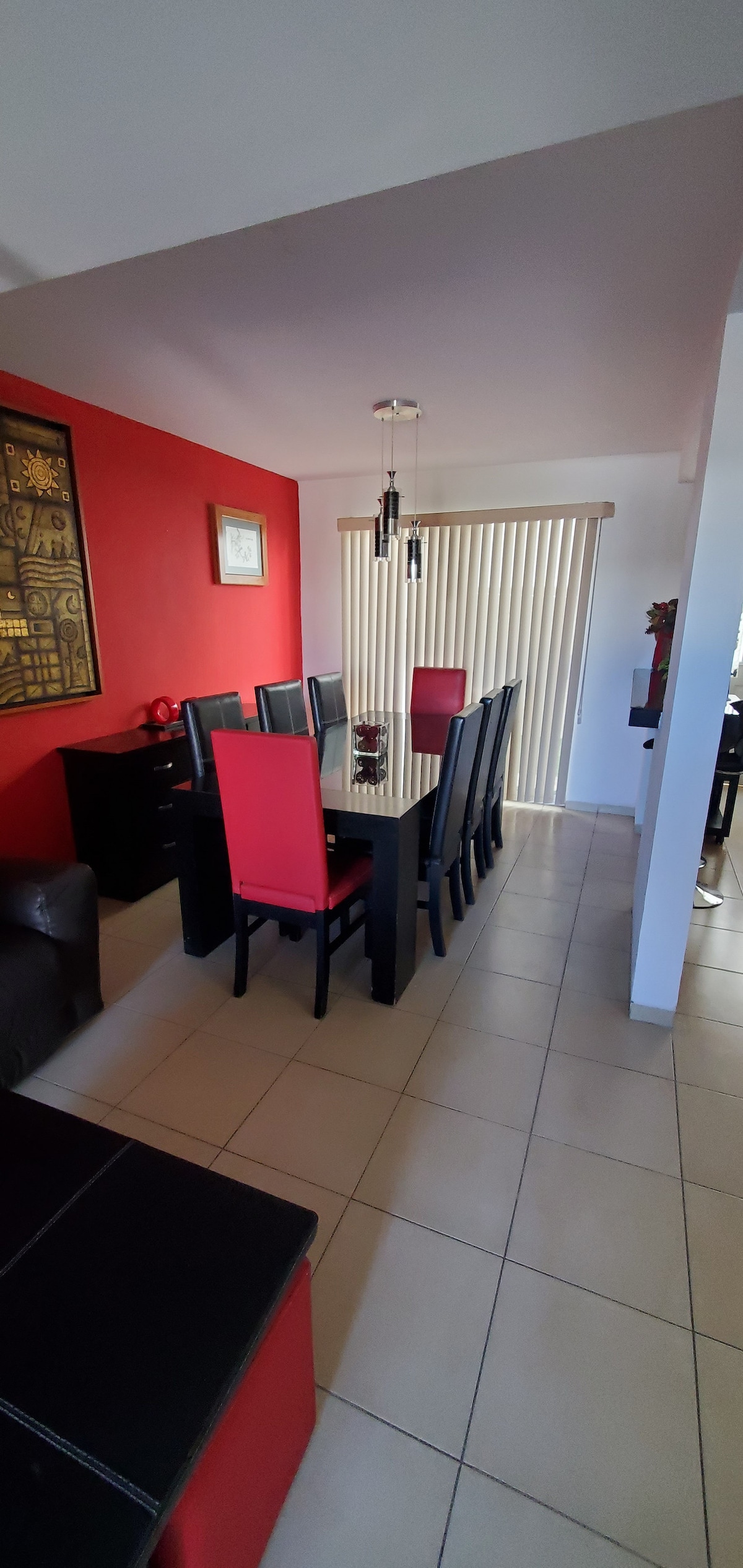A dining area features a black dining table surrounded by eight black and red chairs. A vibrant red accent wall contrasts with the light-colored floor tiles. Natural light enters through vertical blinds covering a window. A dark wooden cabinet is positioned against the wall.