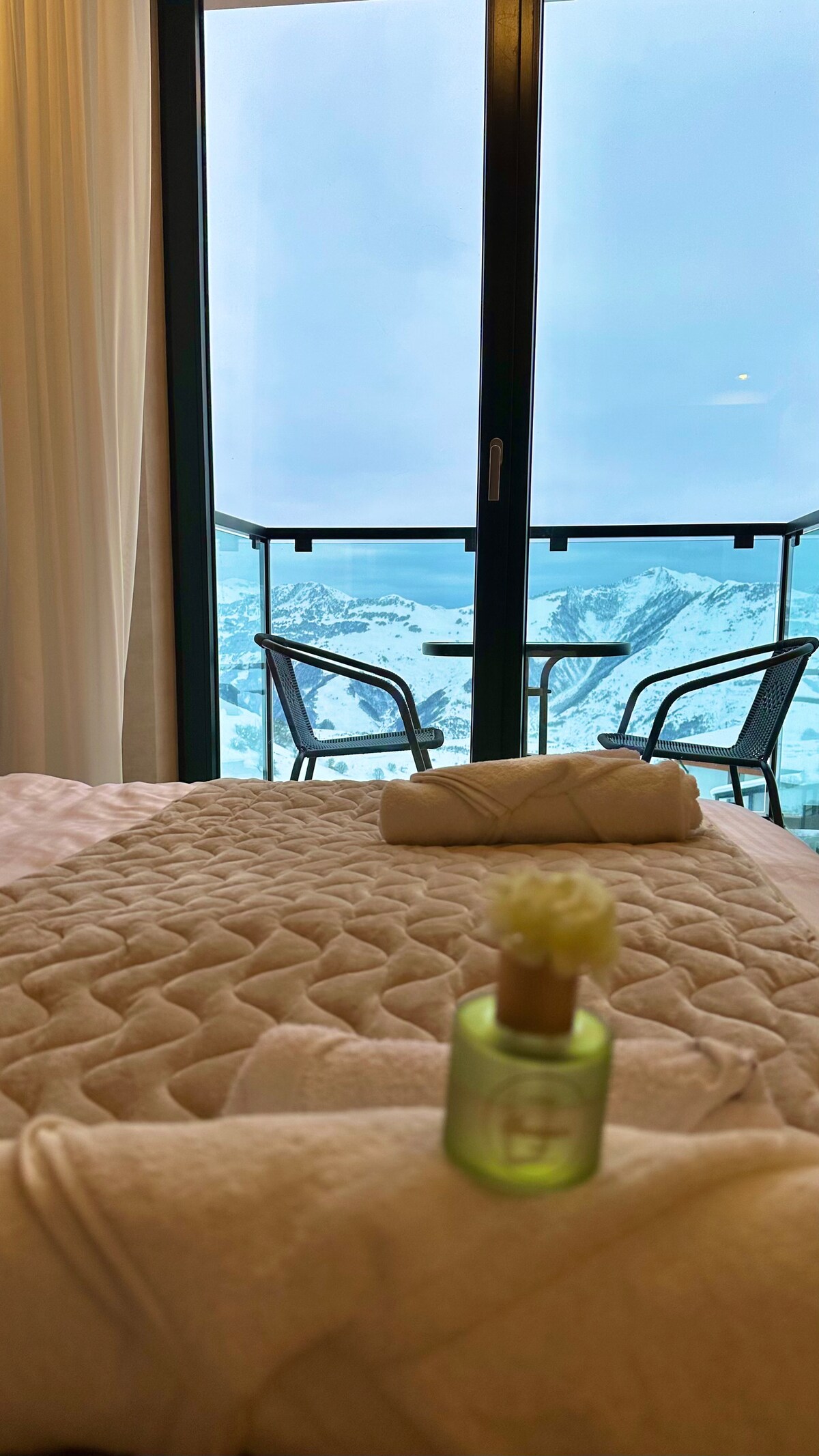 A spacious bed with soft bedding is positioned near a large balcony door. On the bed, rolled towels and a decorative bottle are visible. The balcony offers a view of snow-capped mountains, enhancing the serene ambiance within the apartment.