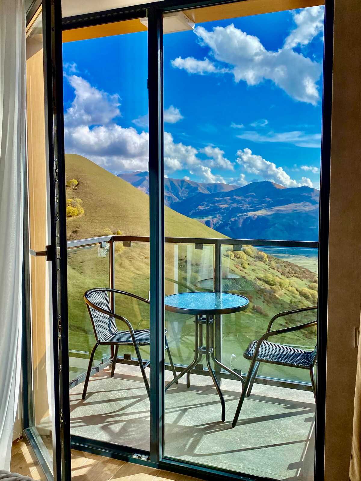 A balcony is seen through glass doors, featuring a round table and two chairs. The backdrop showcases expansive views of green hills under a bright blue sky with scattered clouds.