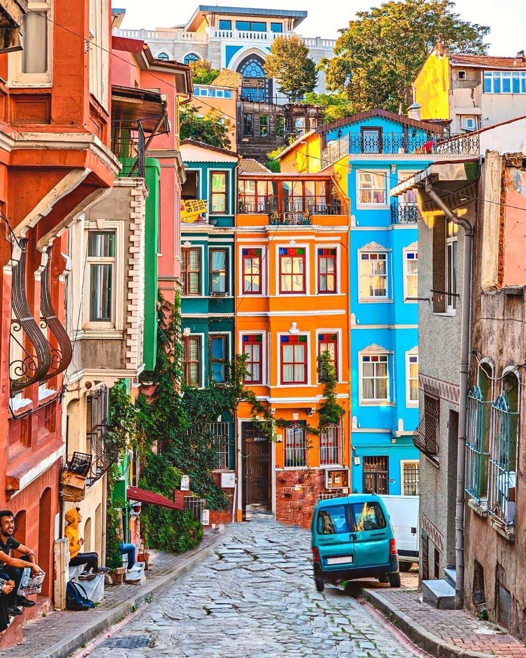 A colorful street in Istanbul is lined with vibrant buildings, showcasing a mix of architectural styles. The cobblestone pathway leads past houses painted in shades of blue, orange, and green. Lush greenery climbs along the walls, adding to the charm of this historic neighborhood.