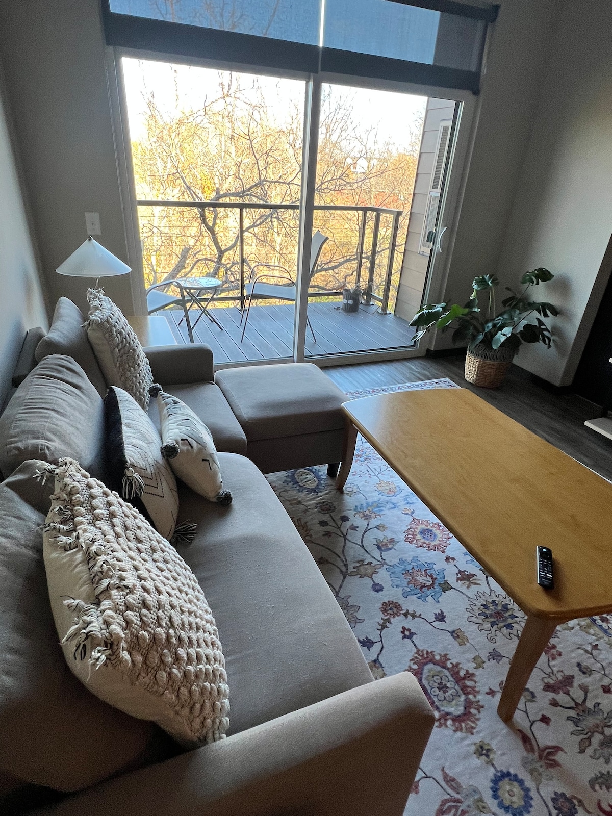 A comfortable seating area features a light brown sofa adorned with decorative pillows. A wooden coffee table sits in front, creating a relaxed space. A glass door opens to a small balcony, with two chairs and greenery visible outside, allowing natural light to fill the room.