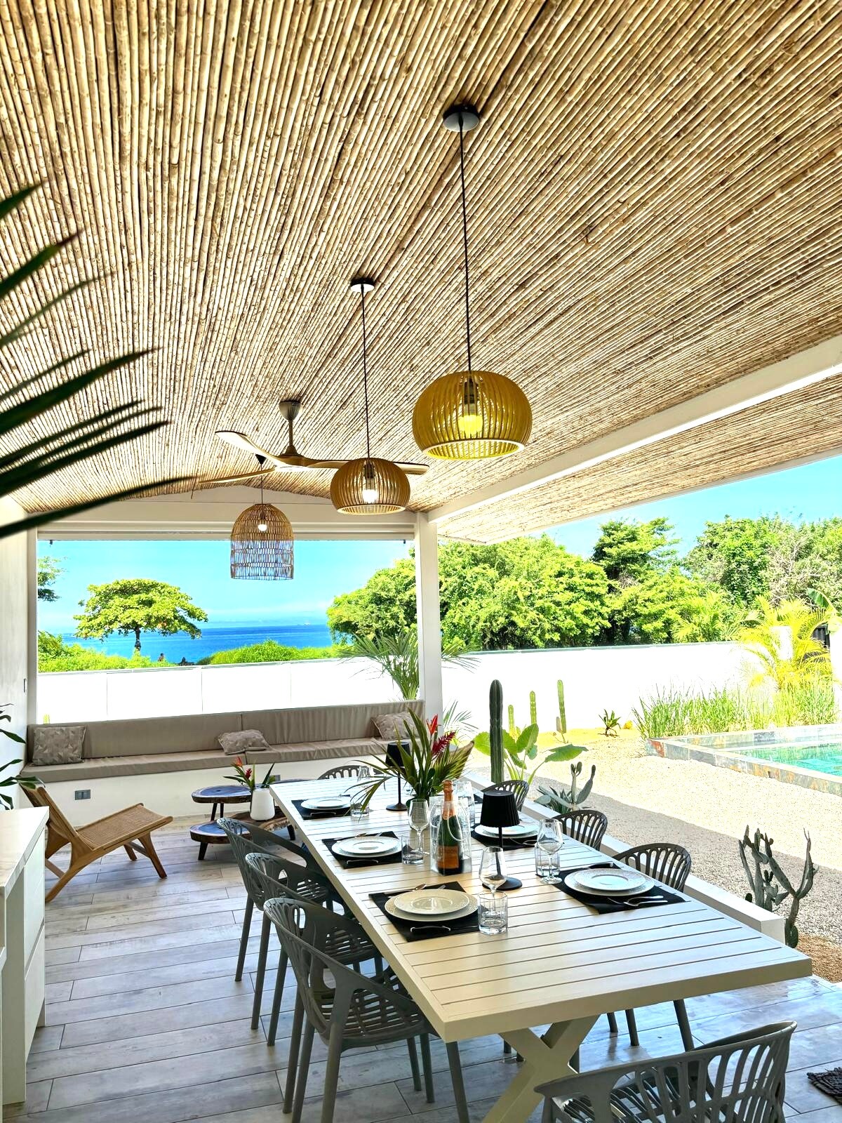 An outdoor dining area is shown, featuring a long white table set for meals. Several gray chairs surround the table, while elegant pendant lights hang from a straw-covered ceiling. A comfortable seating area is visible in the background, along with greenery and the pool.