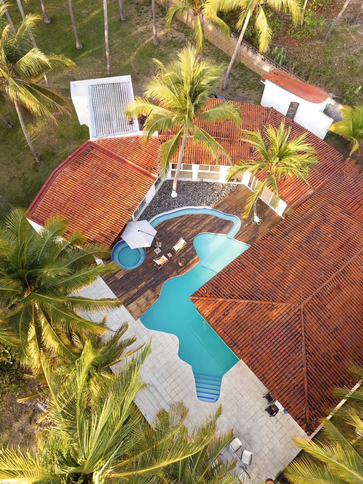 An aerial view of the property showcases a spacious swimming pool surrounded by wooden decking and lounge chairs. The open layout connects the home to lush green areas and vibrant palm trees, while the terracotta roof contrasts beautifully with the surrounding landscape.